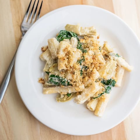 A close-up of the creamy Artichoke Pasta Bake, showcasing wilted spinach and marinated artichoke hearts nestled in penne pasta.